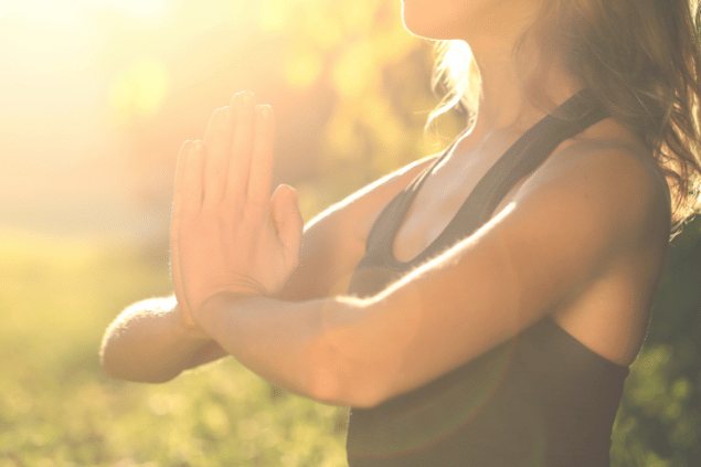 GoodLifeStudio / Getty Images Signature / Canva Uma mulher está com as mãos juntas em forma de oração. Ela medita e sente os raios de sol na pele.