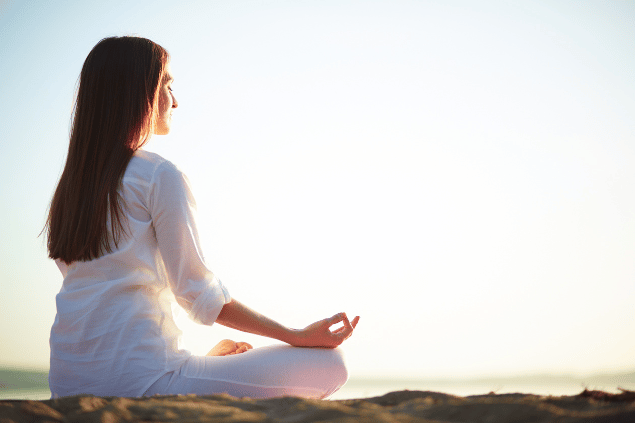 Uma mulher está sentada e meditando em uma praia. Ela sente os raios de sol na pele.