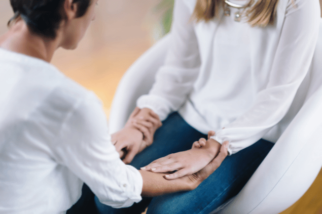 Microgen / Getty Images / Canva Duas mulheres estão de frente de uma para a outra. Elas seguram as mãos, usam roupas brancas e meditam.