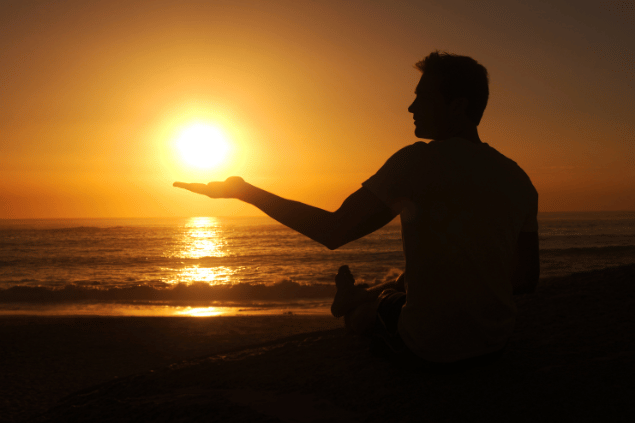 Um homem está sentado de frente para o mar em uma praia. Ele observa e levanta uma mão em direção ao pôr do sol.