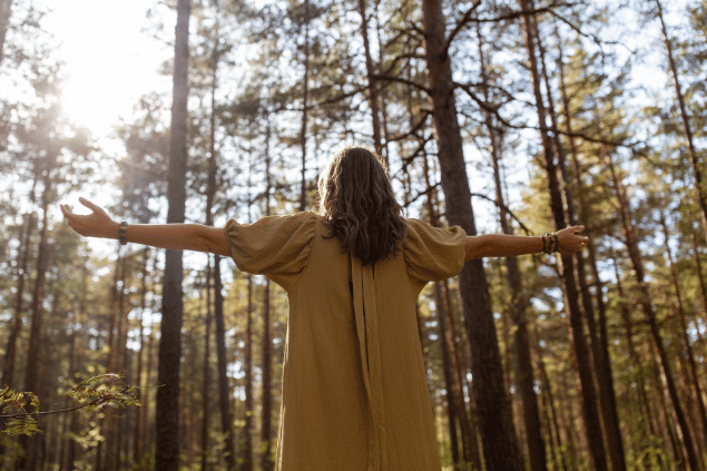 Uma mulher está com os braços abertos em uma floresta. Ela levanta a cabeça e sente os raios de sol na pele.