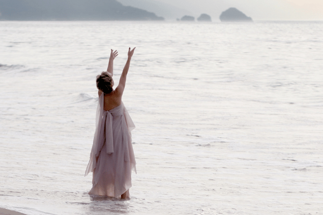 Uma mulher está de pé no raso do mar. Ela levanta a cabeça e os braços e sente a natureza, a espiritualidade.