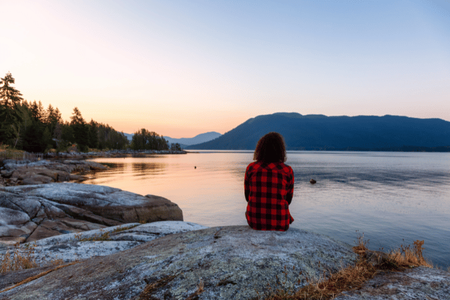 Uma mulher está sentada na frente de um lago em uma floresta. Ela contempla e observa o pôr do sol.