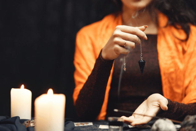Na imagem, as mãos de uma taróloga segurando um pingente com um cristal estão em destaque. Ao lado, há velas e cartas de Tarot.