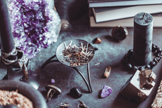 Na imagem, há um recipiente com plantas queimadas em cima de uma mesa. E há uma  vela e pedras também.