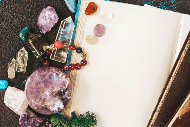 Na imagem, há cristais, plantas e um caderno em cima de uma mesa.