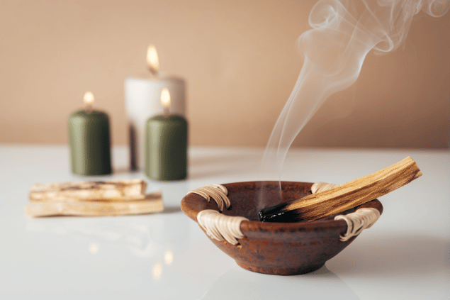 Na imagem, há um recipiente com palo santo e velas acesas em cima de uma mesa.