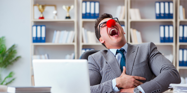 Homem com nariz de palhaço ri. Ele está sentado à mesa de trabalho.