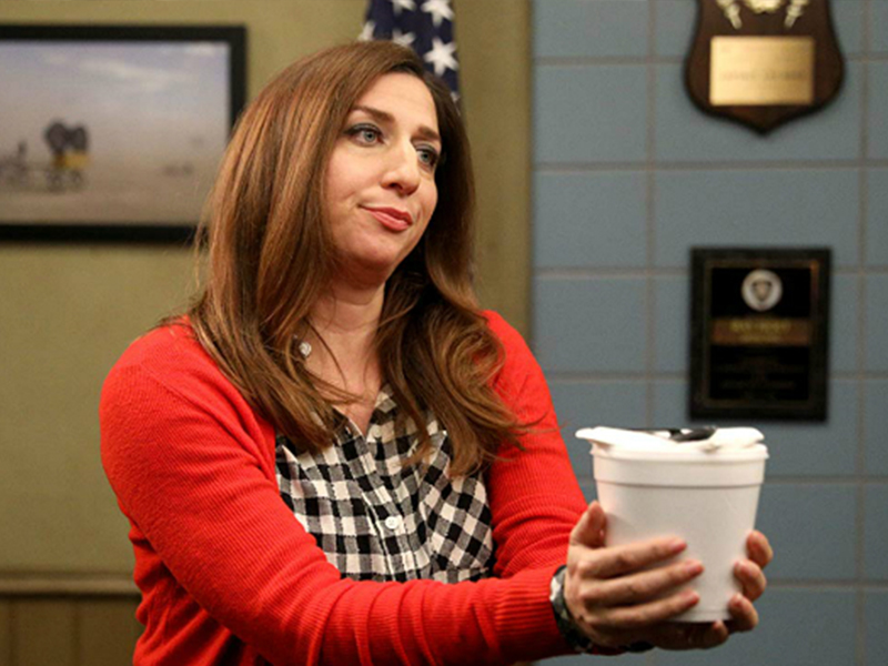 Mulher branca de cabelos médios e ruivos, usando casaco vermelho e camiseta preta e branca, segurando um pote branco.