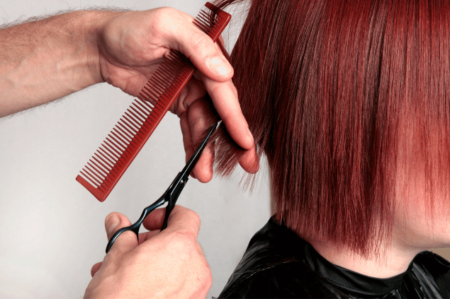 Mão de homem segurando tesoura e pente próximo de um cabelo curto e vermelho. 