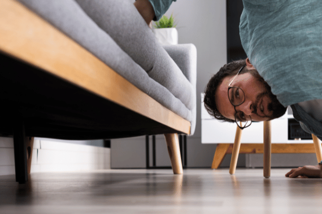 Homem agachado olhando debaixo do sofá enquanto procura por alguma coisa que perdeu. 