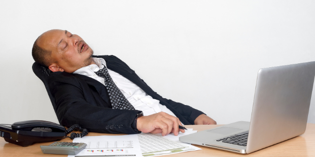Homem sentado em cadeira dormindo. Em sua frente há uma mesa e, sobre ela, um computador, papéis, telefone e calculadora.