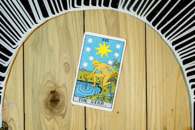 A carta A Estrela está em destaque na imagem sobre uma mesa de madeira. Há outras cartas de Tarot em volta dela.