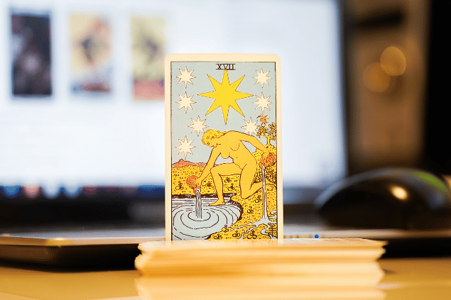 Na imagem, há a carta A Estrela do Tarot na posição normal e em pé. Ao fundo, há um notebook aberto.