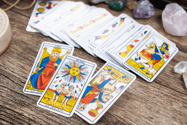 Há três cartas de Tarot em destaque sobre uma mesa de madeira. Em cima, há outras cartas de Tarot e cristais.