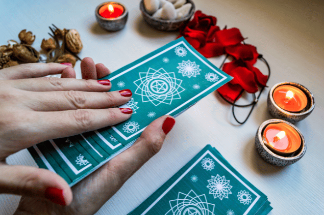 As mãos de uma taróloga segurando algumas cartas de Tarot viradas para baixo estão em destaque na imagem. Algumas velas e flores estão em cima de uma mesa.