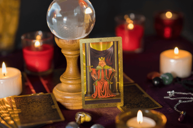 A carta A Justiça está em pé sobre uma mesa. Ela está apoiada em uma bola de cristal. Ao lado, há outras cartas de Tarot, velas e cristais.