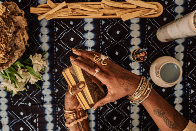 As mãos de uma taróloga segurando um baralho de Tarot estão em destaque na imagem. Há flores e recipientes sobre uma mesa.