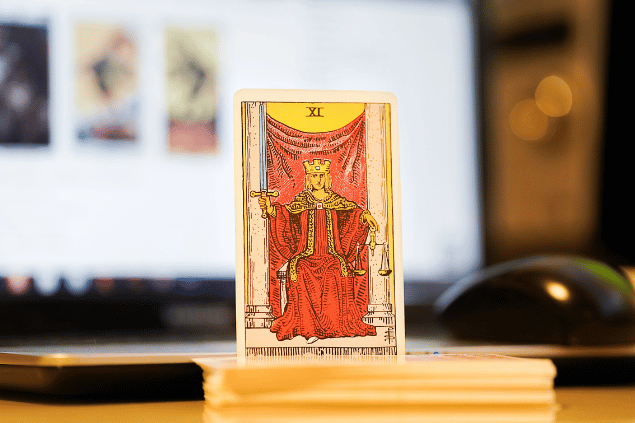 Na imagem, há a carta A Justiça do Tarot na posição normal e em pé. Ao fundo, há um notebook aberto.