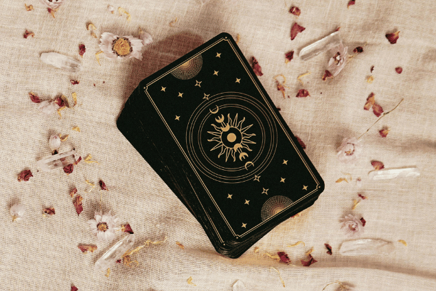 Um baralho de cartas de Tarot na cor preta está em cima de uma superfície. Há algumas flores ao redor dele.