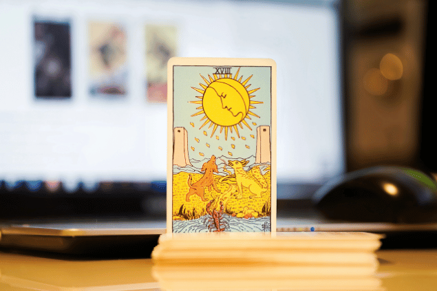 Na imagem, há a carta A Lua do Tarot na posição normal e em pé. Ao fundo, há um notebook aberto.