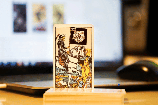 Na imagem, há a carta A Morte do Tarot na posição normal e em pé. Ao fundo, há um notebook aberto.