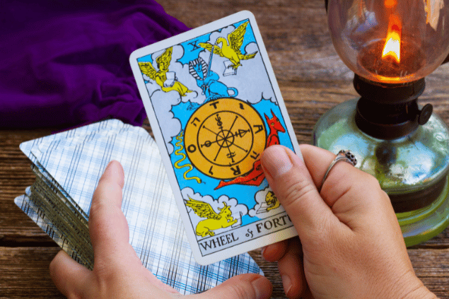 As mãos de uma taróloga seguram um baralho de Tarot e a carta A Roda da Fortuna. Há uma vela ao lado.