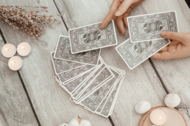 As mãos de uma taróloga segurando três cartas de Tarot viradas para baixo e colocando- as junto com outras cartas sobre uma mesa estão em destaque na imagem. Há velas ao lado.
