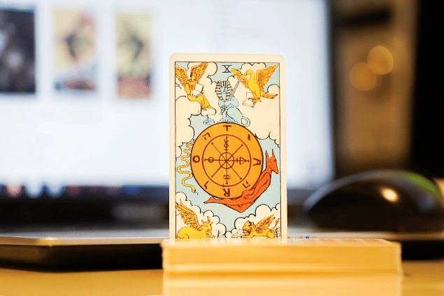 Na imagem, há a carta A Roda da Fortuna do Tarot na posição normal e em pé. Ao fundo, há um notebook aberto.