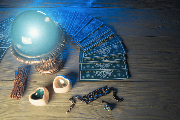 Várias cartas de Tarot viradas para baixo estão dispostas sobre uma mesa. Há uma bola de cristal e velas ao lado.