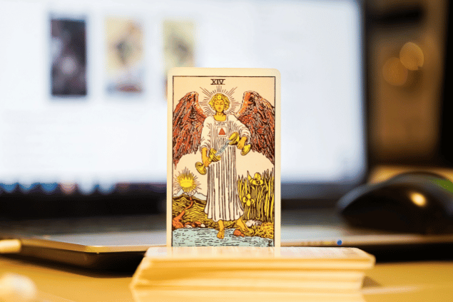 Na imagem, há a carta A Temperança do Tarot na posição normal e em pé. Ao fundo, há um notebook aberto.