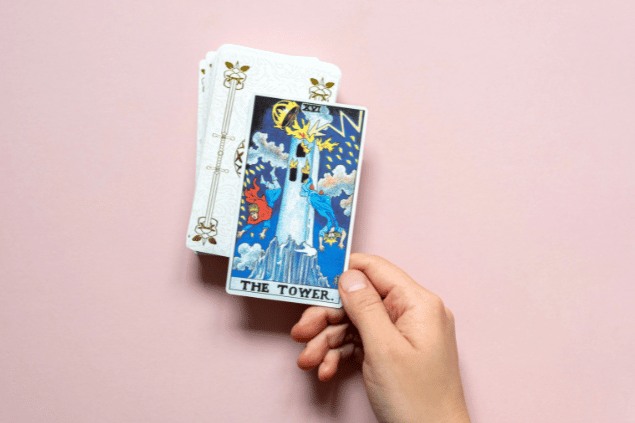 A mão de uma pessoa segura a carta A Torre do Tarot que está em destaque na imagem. Embaixo dela, há um baralho com outras cartas viradas para baixo.