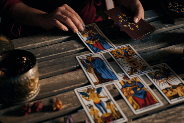Seis cartas de Tarot estão dispostas sobre uma mesa de madeira. Uma taróloga segura um baralho com outras cartas na mão.