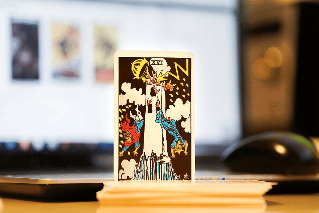 Na imagem, há a carta A Torre do Tarot na posição normal e em pé. Ao fundo, há um notebook aberto.