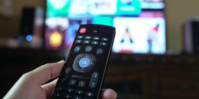 sinseeho / Canva Pessoa segura controle remoto e o aponta para televisão ligada.
