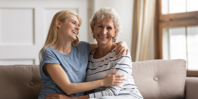 Duas mulheres, uma mais jovem e outra mais velha, sentadas lado a lado em um sofá cor creme. Elas estão abraçadas e sorriem. O cenário é de sala de estar.