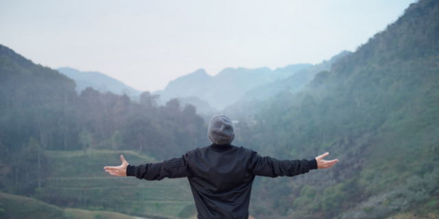 Homem com os braços erguidos e abertos em cenário externo de montanhas.