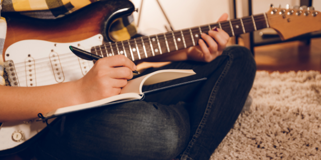 Mulher segura guitarra pelo braço. Sobre sua perna, há um caderno no qual escreve. Ela está sentado no chão.