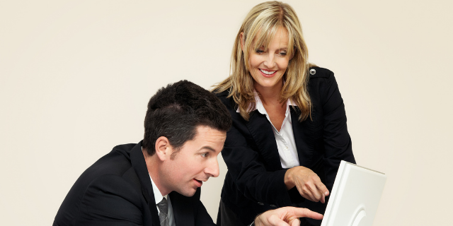 Homem sentado à mesa aponta para computador. Uma mulher está de pé ao seu lado e faz o mesmo gesto. 