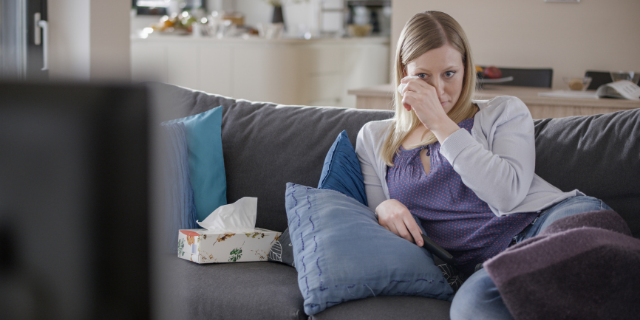 Mulher chora em frente à TV e enxuga o rosto com lenço de papel.