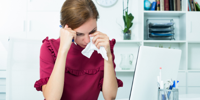 Mulher sentada à mesa em um escritório chora. Ela enxuga o rosto com um lenço de papel.