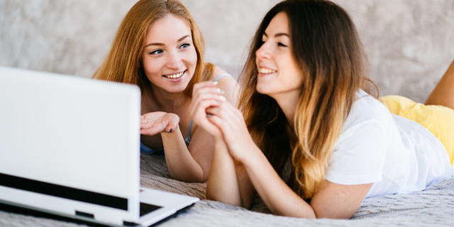 Duas mulheres deitadas de bruços em cama. Ao lado delas, há um computador. Ambas sorriem.