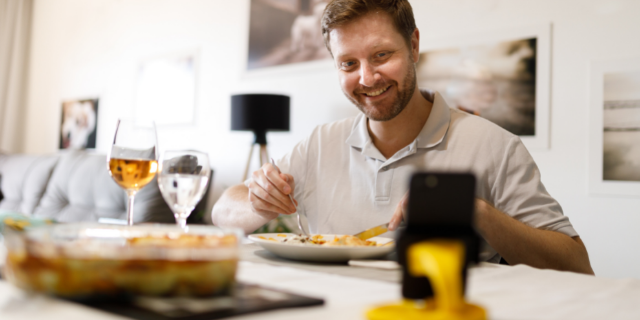 Capuski / Canva Homem sentado à mesa tem uma refeição em frente do celular que está sobre a mesa. Ele sorri.