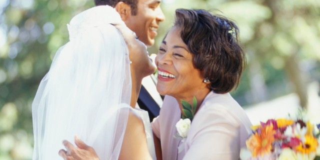 Duas mulheres, sogra e nora, abraçam-se em cerimônia de casamento. Ao fundo, há um homem vestido socialmente. O cenário é externo e arborizado.