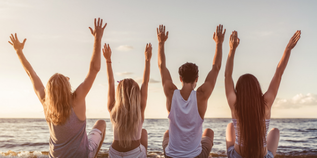 Quatro pessoas sentadas à beira do mar. Eles estão com os braços erguidos.
