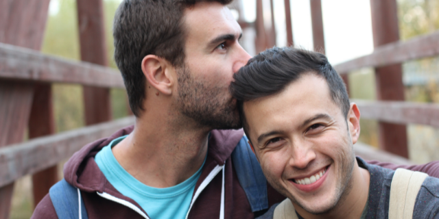 Homem abraça outro ao seu lado e beija sua cabeça.