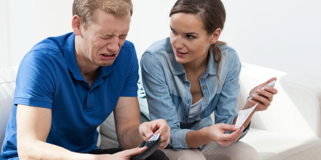 Homem e mulher sentados em sofá branco. Ele chora e segura uma carteira aberta, ela o olha enquanto segura um papel e uma calculadora.