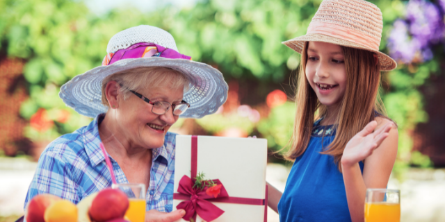 Avó e neta em ambiente externo. A primeira entrega à segunda um cartão com flores. Ambas sorriem.