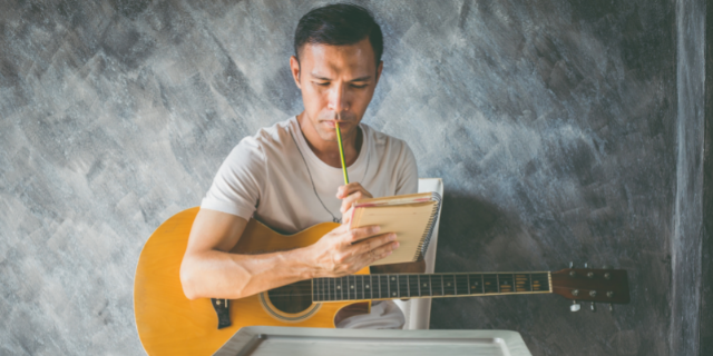 Homem com um violão próximo do corpo. Ele segura um caderno e um lápis próximo da boca.