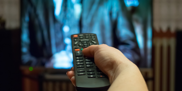 Mão segura controle remoto em frente de uma televisão.